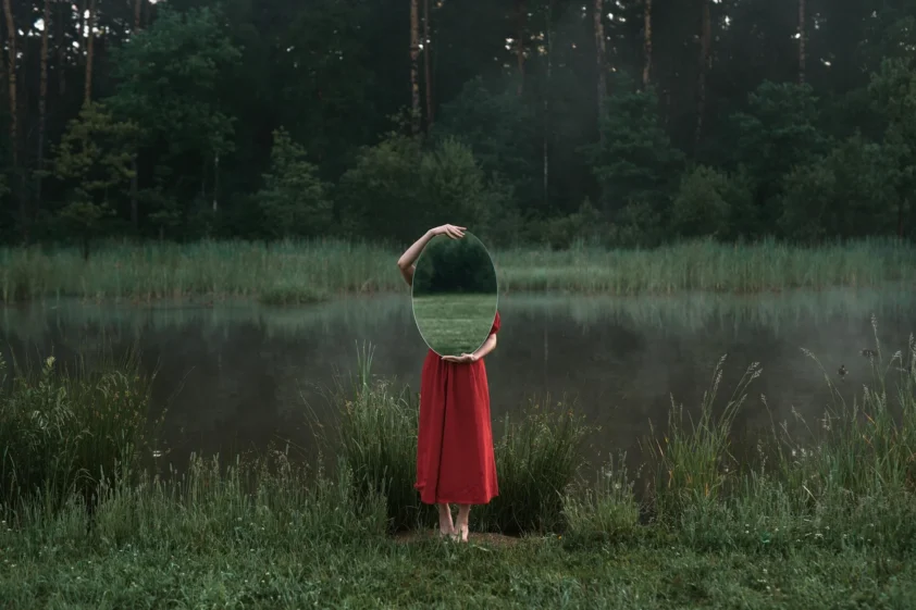 a woman in red dress standing on green grass while holding a mirror